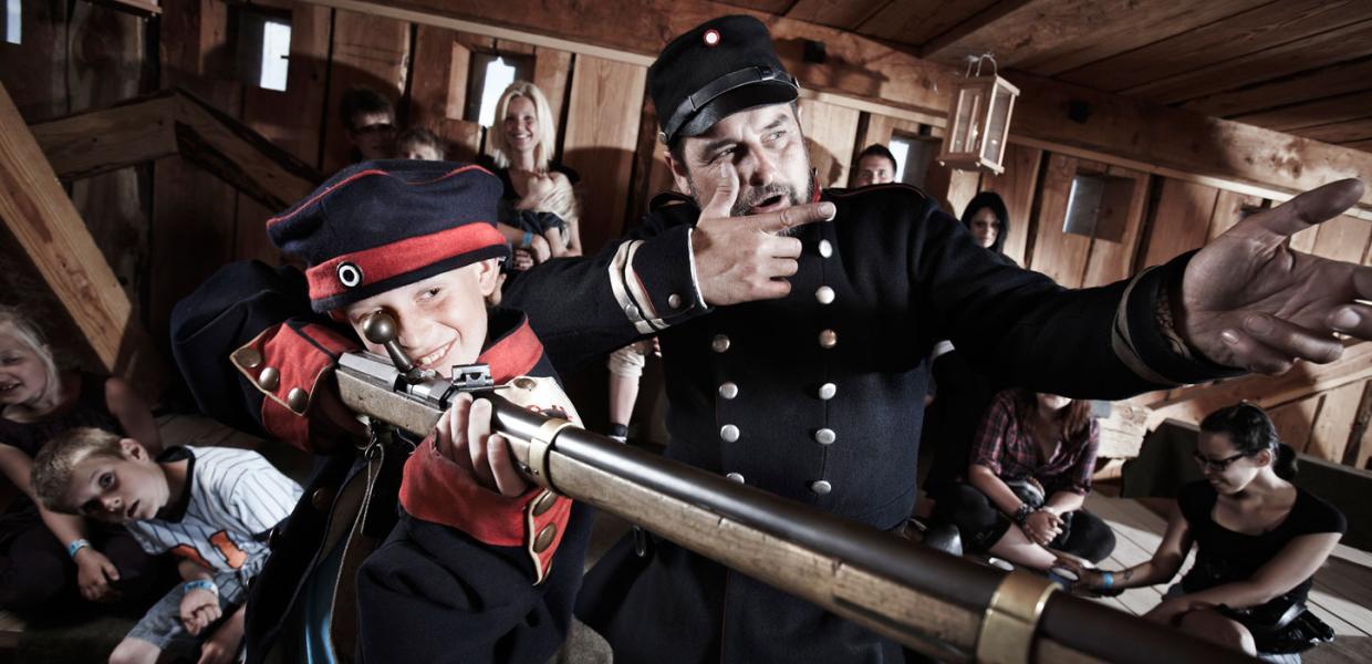 Medarbejder (formidler) og barn i uniform skyder med trægevær på Historiecenter Dybbøl Banke
